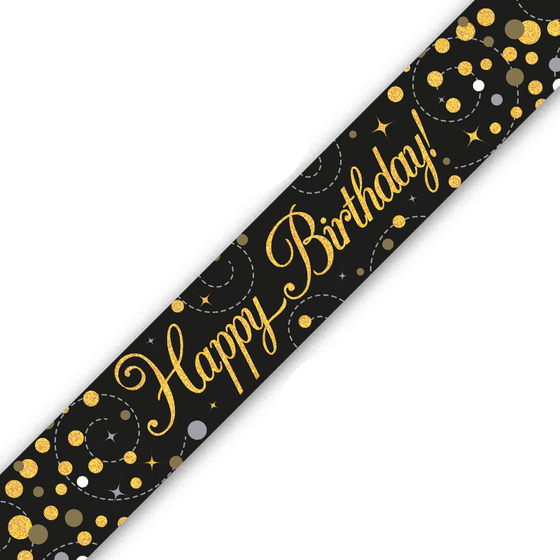 A black birthday banner or sash printed with 'Happy Birthday!' in gold cursive script, decorated with gold, silver, and white confetti dots and stars, shown diagonally on a white background.