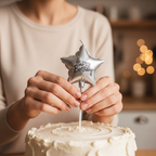 Hands placing a silver foil birthday candle onto a white birthday cake in a warm kitchen setting