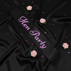 Black hen party sash with pink writing displayed on black satin with pink roses and confetti