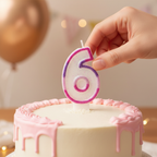 Hand placing pink glitter number 6 birthday candle onto white birthday cake, candid lifestyle moment with bokeh background