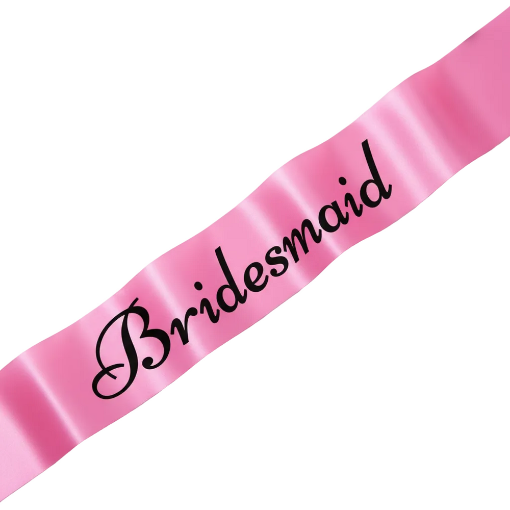 Pink satin bridesmaid sash with bold black writing, elegantly displayed on a blush marble surface with white rose petals