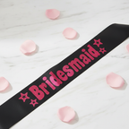 Black satin bridesmaid sash with hot pink foil letters displayed on white marble with pink rose petals – elegant product shot