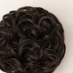 Extreme macro close-up of dark brown curly hair bun 2 Nigella showing individual curl texture, warm espresso mocha undertones and strand craftsmanship
