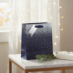 Navy ombre gift bag with silver ribbon bow styled on a marble surface with eucalyptus and fairy lights in a home interior setting
