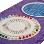 Extreme macro close-up of assorted coloured pin heads in white circular tray showing vivid red, yellow, green and silver needle detail
