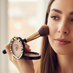 Woman applying W7 The Bronzer Matte Bronzing Powder Compact to cheekbones with fluffy brush, candid beauty moment with warm bokeh background