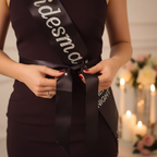 Close up of hands tying black satin diamante bridesmaid sash into bow at waist with wedding candles in background