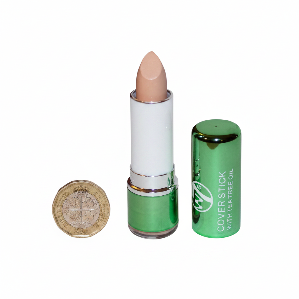 W7 Tea Tree Concealer Stick Light/Medium flat lay beside £1 coin for size reference, white background with soft overhead lighting showing metallic green casing and light peachy-beige bullet