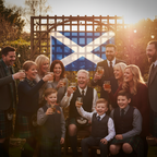 Joyful group celebrating Burns Night with Scotland flag as backdrop, golden hour warm light and glasses raised