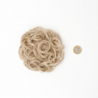 Flat lay of warm blonde curly messy hair bun beside a £1 coin showing product size, clean white background with soft overhead lighting