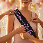 Close-up of black hen party sash with pink Mother of the Groom writing being adjusted on woman's shoulder at celebration venue