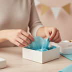 Woman's hands placing light blue tissue paper into a white gift box, candid gift wrapping moment with warm bokeh background
