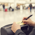 Hands writing name and address on a pale yellow manilla tie-on luggage label attached to a suitcase handle, candid travel preparation moment