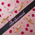 Hen party flat lay featuring black bridesmaid sash with pink writing surrounded by tiara, prosecco, and confetti