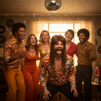 Group of friends celebrating at 70s fancy dress party, one wearing brown mullet wig and moustache
