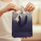 Hands placing silver tissue paper into a navy ombre gift bag with metallic silver dot pattern and navy cord handles