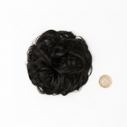 Flat lay of dark brown curly messy hair bun 1B Queen of the Night beside a £1 coin showing product size, clean white background with soft overhead lighting