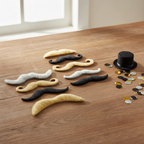Six assorted fancy dress moustaches in blonde, black and grey arranged on a rustic table with party props and natural window light