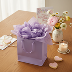 Gift bag overflowing with lilac tissue paper surrounded by spring flowers and Mother's Day gifts