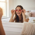 Young woman trying on hippy glasses with pink lenses in front of a mirror, smiling in a bright cosy bedroom