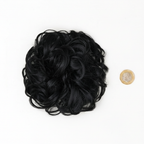 Flat lay of jet black curly messy hair bun beside a £1 coin showing product size, clean white background with soft overhead lighting