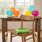 Neon green whoopee cushion on a wooden chair at a party table with colourful balloons, natural window light