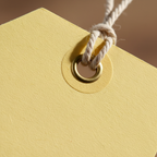 Extreme macro close-up of a manilla luggage label showing pale yellow card texture, reinforced metal eyelet and natural cream twisted cotton twine