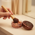 Woman swirling makeup brush through Body Collection Bronzing Pearls jar — candid lifestyle shot with warm bokeh background