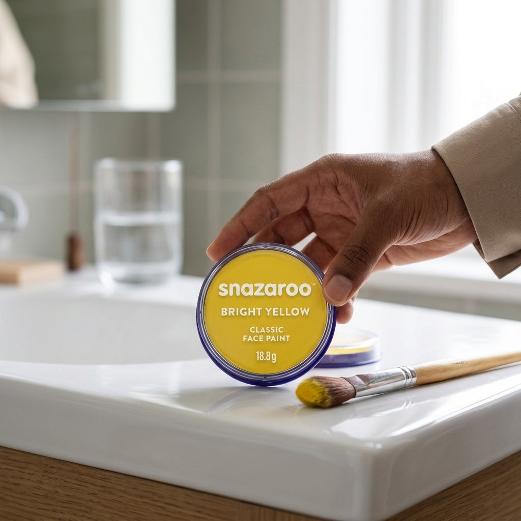 A hand holding a round tin of Snazaroo Bright Yellow Classic Face Paint over a white sink, with a paintbrush nearby and a softly blurred bathroom background.