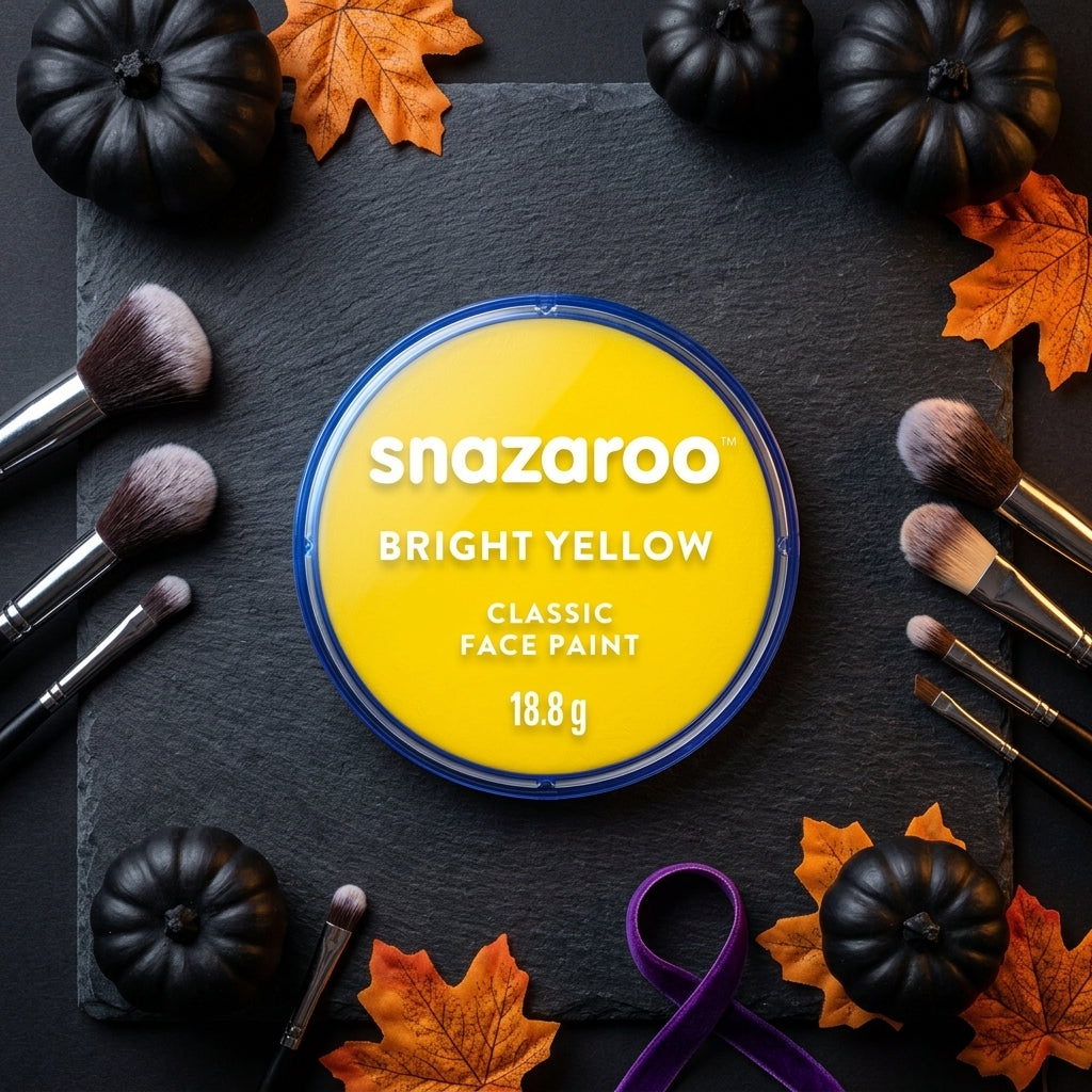 A round tin of Snazaroo Bright Yellow Classic Face Paint is centered on a dark slate background, surrounded by decorative black pumpkins, orange autumn leaves, makeup brushes, and a purple ribbon.