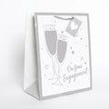 A white and silver gift bag with a champagne-themed design, featuring two champagne glasses, small hearts, and the text 'On Your Engagement'.