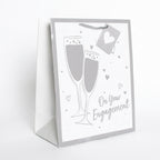 A white and silver gift bag with a champagne-themed design, featuring two champagne glasses, small hearts, and the text 'On Your Engagement'.