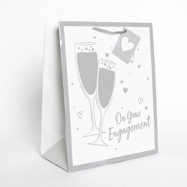 A white and silver gift bag with a champagne-themed design, featuring two champagne glasses, small hearts, and the text 'On Your Engagement'.